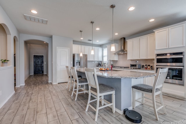 kitchen featuring an island with sink, wall chimney exhaust hood, light stone countertops, stainless steel appliances, and decorative light fixtures