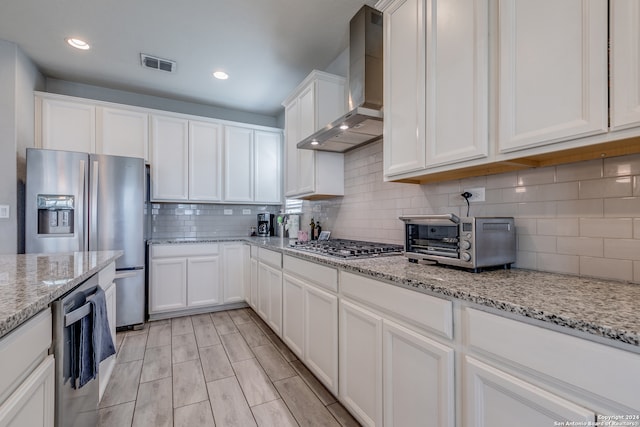 kitchen featuring wall chimney exhaust hood, backsplash, white cabinetry, appliances with stainless steel finishes, and light stone countertops