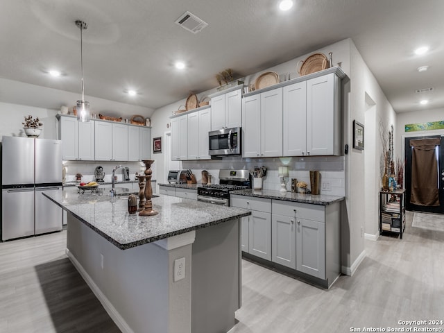 kitchen featuring light stone counters, hanging light fixtures, a center island with sink, stainless steel appliances, and light wood-type flooring