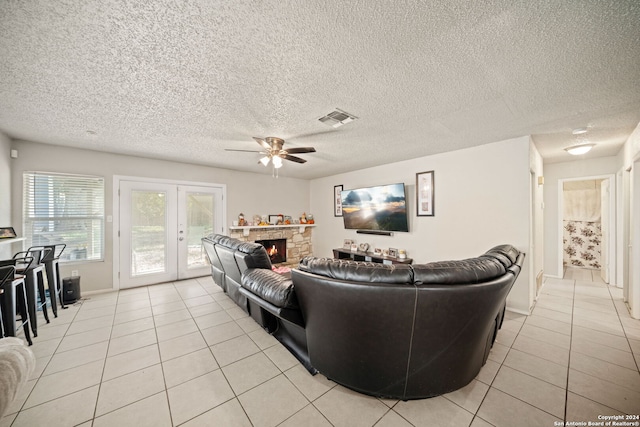 tiled living room with ceiling fan, a textured ceiling, french doors, and a stone fireplace