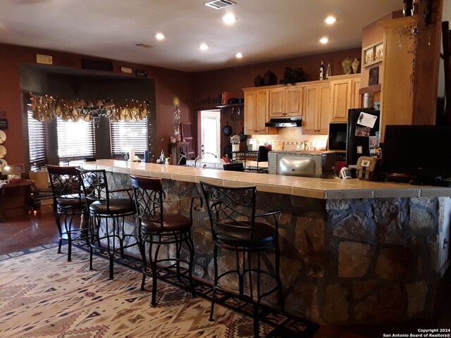 kitchen with backsplash, kitchen peninsula, tile countertops, a kitchen bar, and light brown cabinetry