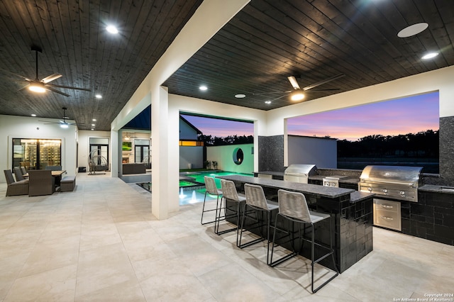 patio terrace at dusk with area for grilling, ceiling fan, a bar, and an outdoor kitchen