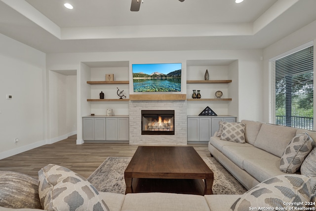 living room with light hardwood / wood-style floors, a tile fireplace, a raised ceiling, ceiling fan, and built in shelves