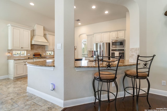 kitchen with white cabinets, kitchen peninsula, stone counters, stainless steel appliances, and custom range hood