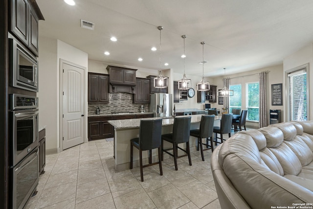 kitchen with dark brown cabinetry, hanging light fixtures, tasteful backsplash, a center island with sink, and stainless steel appliances