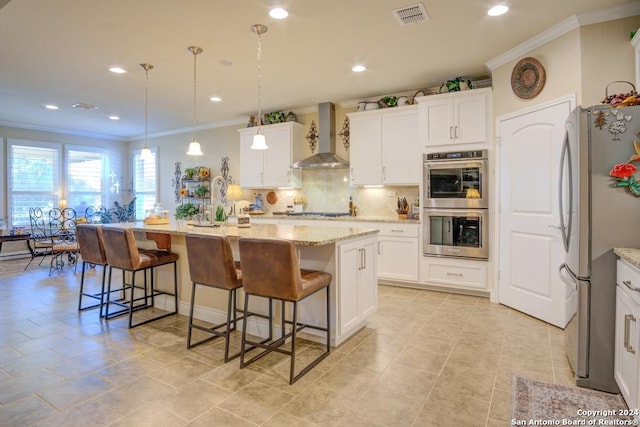 kitchen with an island with sink, stainless steel appliances, hanging light fixtures, and wall chimney range hood