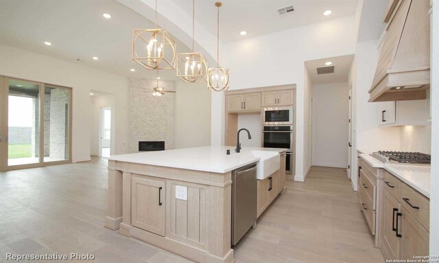 kitchen featuring premium range hood, a kitchen island with sink, appliances with stainless steel finishes, decorative light fixtures, and light brown cabinets