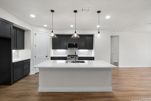 kitchen featuring a kitchen island with sink, tasteful backsplash, hanging light fixtures, stainless steel appliances, and hardwood / wood-style floors