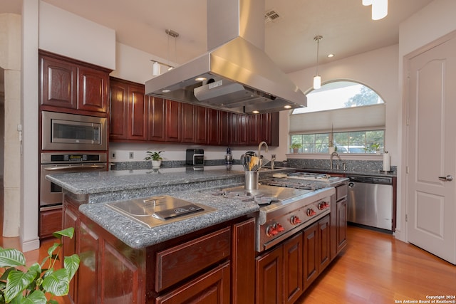 kitchen with appliances with stainless steel finishes, island exhaust hood, hanging light fixtures, light hardwood / wood-style floors, and a center island with sink