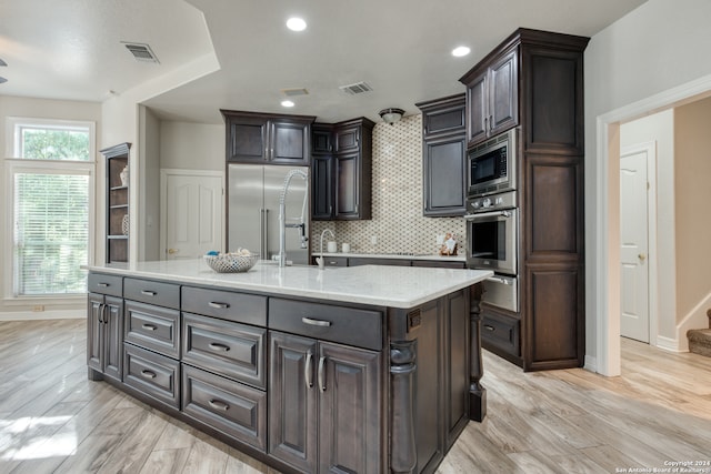 kitchen featuring a center island with sink, dark brown cabinetry, built in appliances, and light wood-type flooring