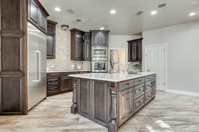 kitchen featuring built in appliances, a center island with sink, sink, dark brown cabinetry, and tasteful backsplash