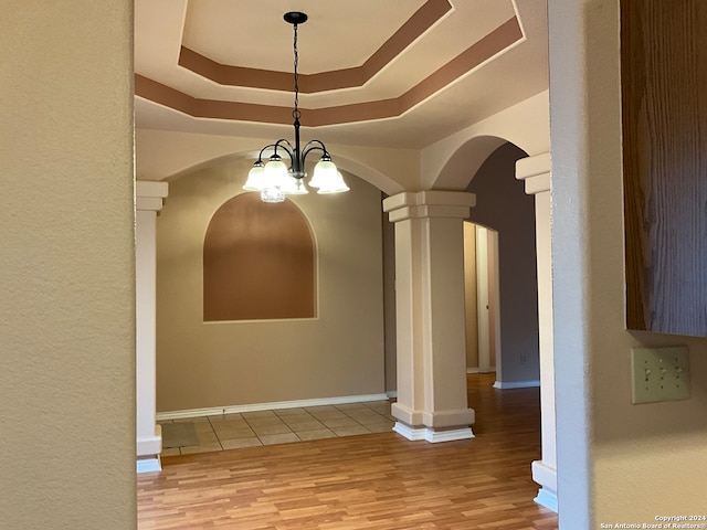 unfurnished dining area featuring a notable chandelier, a tray ceiling, wood-type flooring, and decorative columns