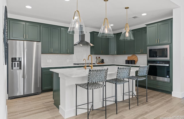 kitchen featuring hanging light fixtures, a center island with sink, wall chimney exhaust hood, stainless steel appliances, and light wood-type flooring