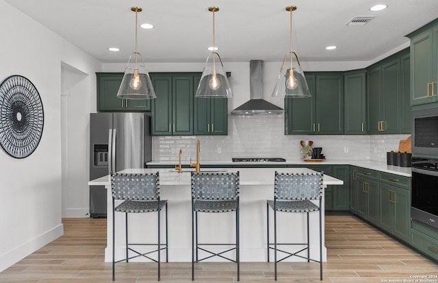 kitchen with light hardwood / wood-style flooring, a center island with sink, appliances with stainless steel finishes, and wall chimney range hood