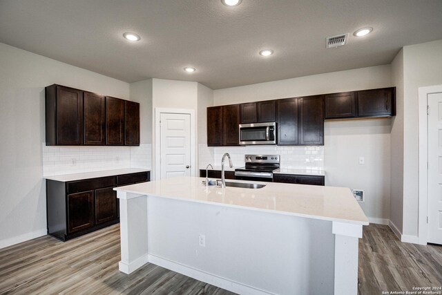 kitchen with a center island with sink, light hardwood / wood-style flooring, decorative backsplash, dark brown cabinetry, and stainless steel appliances