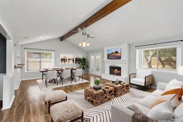 living room featuring a brick fireplace, ceiling fan, hardwood / wood-style flooring, vaulted ceiling with beams, and a textured ceiling