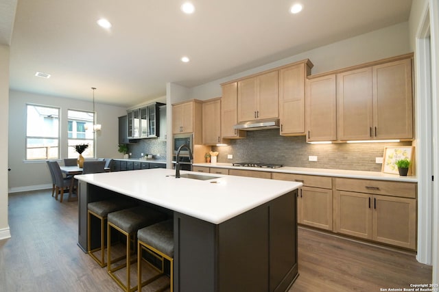 kitchen with light brown cabinetry, sink, hanging light fixtures, stainless steel appliances, and a kitchen island with sink