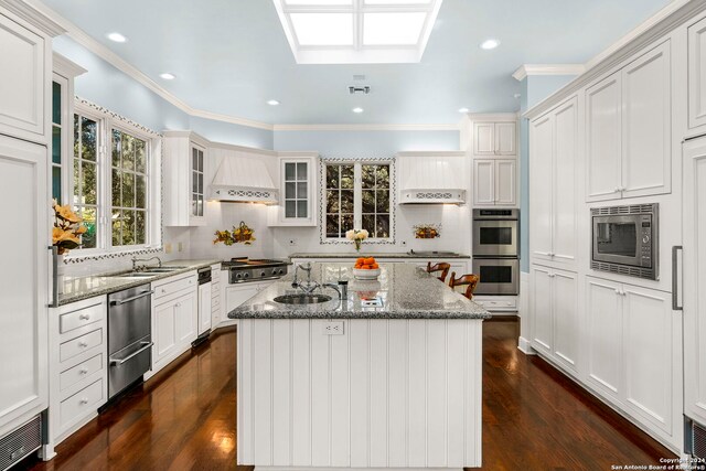 kitchen with an island with sink, premium range hood, light stone countertops, sink, and stainless steel appliances