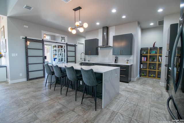 kitchen featuring wall chimney range hood, tasteful backsplash, a center island with sink, decorative light fixtures, and light stone countertops