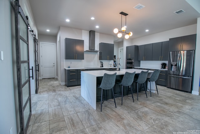 kitchen featuring a barn door, wall chimney exhaust hood, a kitchen island with sink, decorative light fixtures, and stainless steel appliances