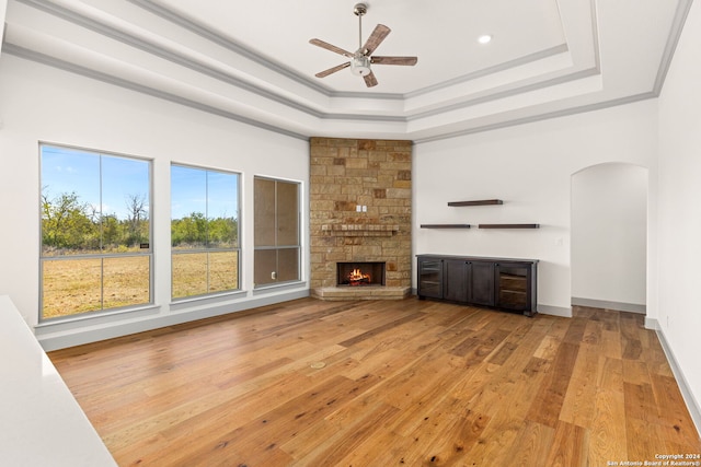 unfurnished living room featuring ceiling fan, a stone fireplace, a raised ceiling, light hardwood / wood-style floors, and crown molding