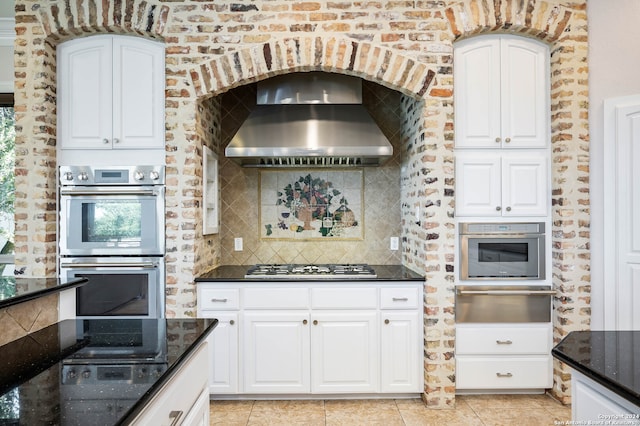 kitchen featuring wall chimney exhaust hood, tasteful backsplash, appliances with stainless steel finishes, light tile patterned floors, and white cabinetry