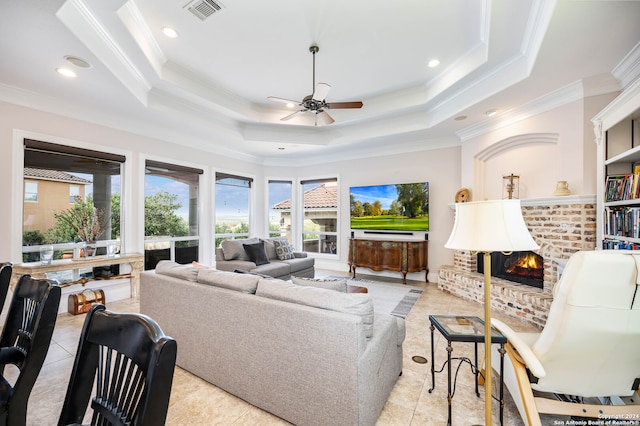 living room featuring a brick fireplace, ceiling fan, light tile patterned flooring, a tray ceiling, and ornamental molding