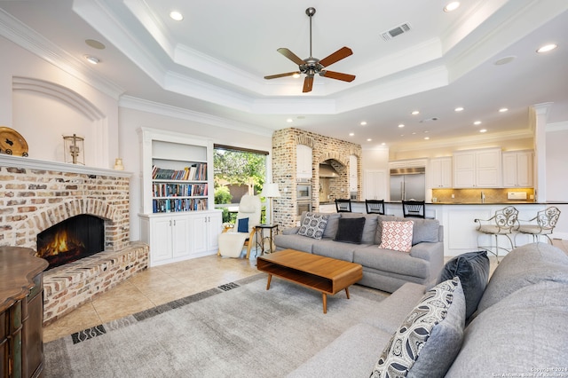 living room with ornamental molding, a brick fireplace, a tray ceiling, and light tile patterned floors