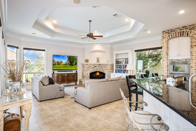 living room featuring ceiling fan, a raised ceiling, crown molding, a fireplace, and light tile patterned floors