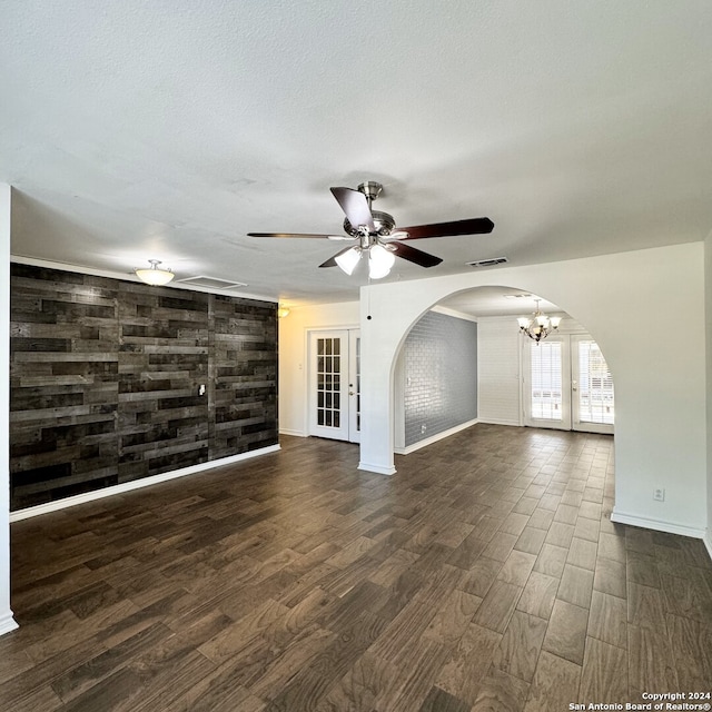 unfurnished living room with french doors, ceiling fan with notable chandelier, wood walls, and dark wood-type flooring