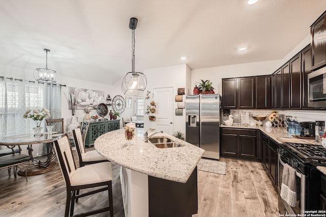 kitchen with light wood-type flooring, a kitchen island with sink, sink, appliances with stainless steel finishes, and decorative light fixtures