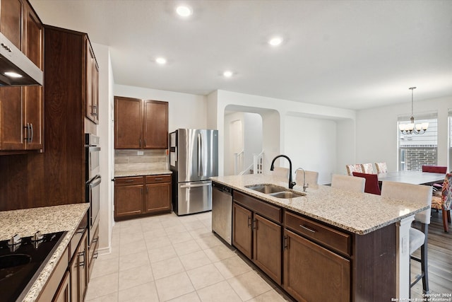 kitchen with a kitchen island with sink, an inviting chandelier, sink, stainless steel appliances, and light stone countertops