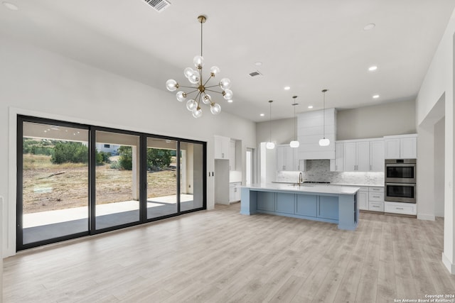 kitchen featuring white cabinets, decorative light fixtures, a center island with sink, double oven, and light hardwood / wood-style floors