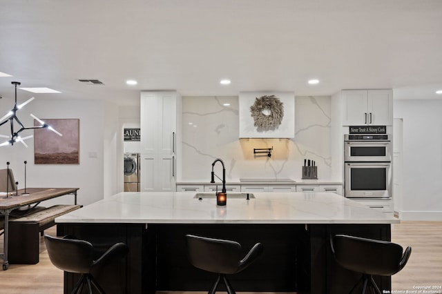 kitchen featuring light wood-type flooring, stainless steel double oven, sink, an island with sink, and white cabinetry