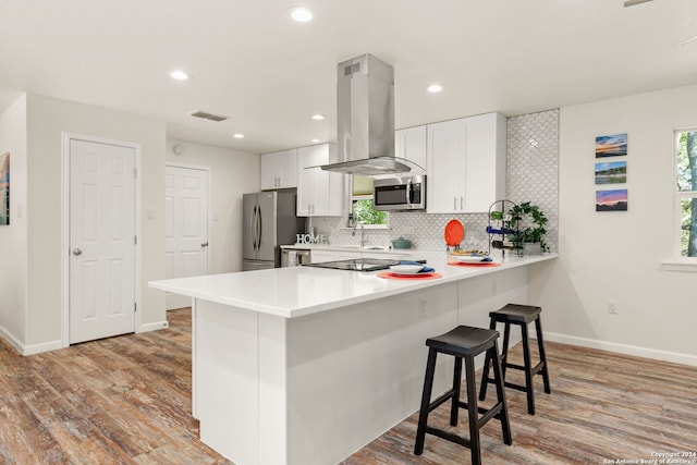 kitchen with light wood-type flooring, kitchen peninsula, island range hood, white cabinetry, and appliances with stainless steel finishes