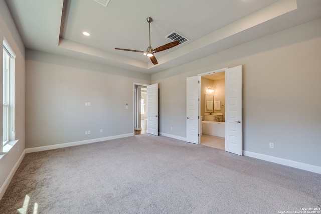 unfurnished bedroom with light carpet, a tray ceiling, ceiling fan, and ensuite bathroom