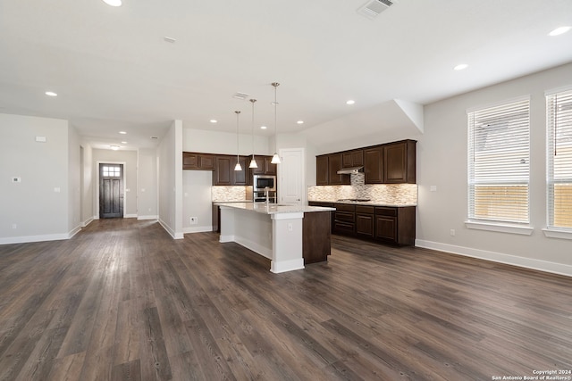 kitchen featuring an island with sink, hanging light fixtures, backsplash, stainless steel appliances, and dark hardwood / wood-style floors