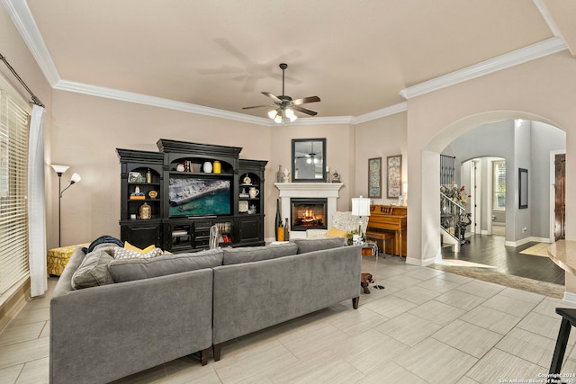 living room featuring arched walkways, a glass covered fireplace, a ceiling fan, baseboards, and ornamental molding