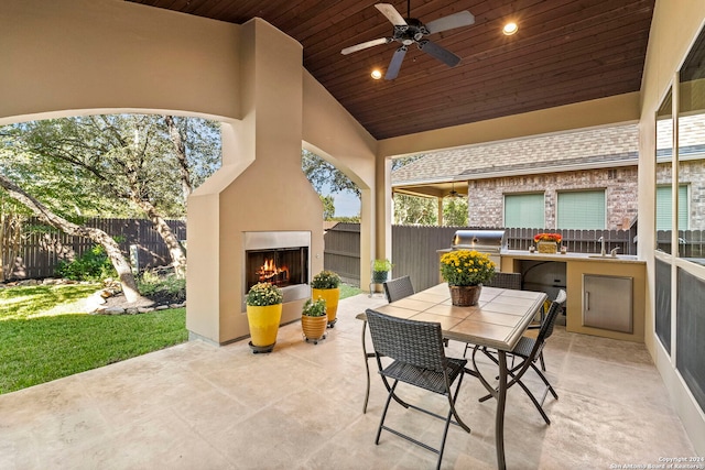 view of patio / terrace featuring a ceiling fan, a warm lit fireplace, a fenced backyard, an outdoor kitchen, and a grill