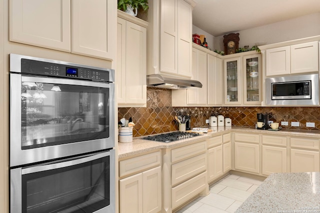 kitchen featuring light stone countertops, stainless steel appliances, tasteful backsplash, under cabinet range hood, and glass insert cabinets