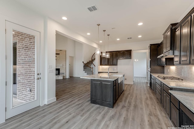 kitchen with pendant lighting, a fireplace, stainless steel gas cooktop, dark brown cabinets, and a center island with sink