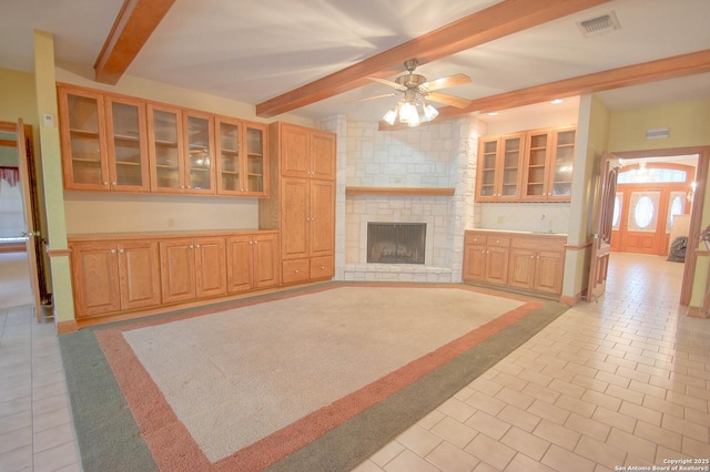 unfurnished living room with ceiling fan, light tile patterned floors, beam ceiling, and a stone fireplace