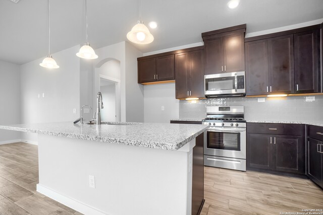 kitchen featuring an island with sink, tasteful backsplash, decorative light fixtures, stainless steel appliances, and light wood-type flooring