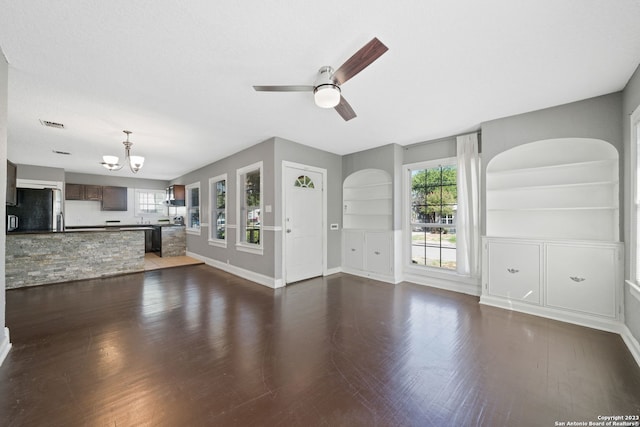 unfurnished living room with ceiling fan with notable chandelier, dark hardwood / wood-style floors, built in shelves, and a healthy amount of sunlight