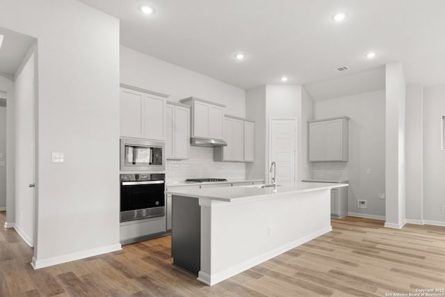 kitchen featuring decorative backsplash, appliances with stainless steel finishes, light hardwood / wood-style floors, and an island with sink