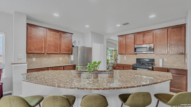 kitchen featuring stone countertops, decorative backsplash, a kitchen island with sink, and stainless steel appliances