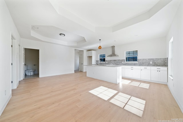 kitchen featuring pendant lighting, wall chimney range hood, an island with sink, a tray ceiling, and white cabinetry