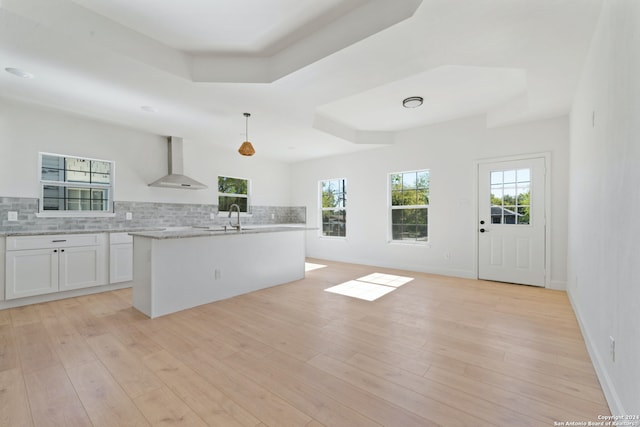 kitchen featuring wall chimney exhaust hood, a raised ceiling, pendant lighting, white cabinets, and an island with sink