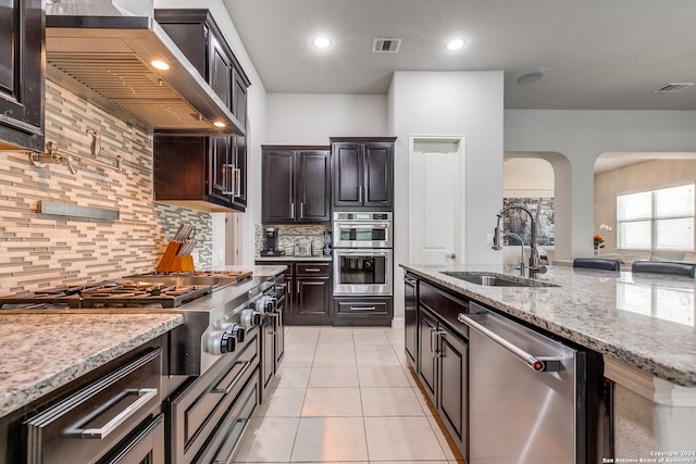 kitchen with wall chimney range hood, sink, light stone counters, stainless steel appliances, and backsplash