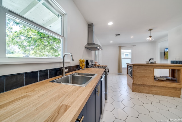 kitchen with wall chimney exhaust hood, wooden counters, decorative light fixtures, sink, and appliances with stainless steel finishes